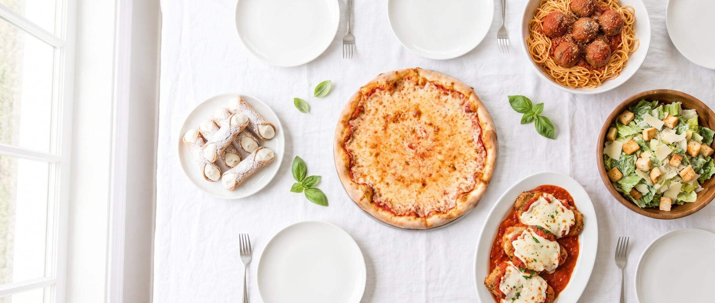 A family-style spread on white linen: pizza, spaghetti and meatballs, Caesar salad, chicken parmigiana and cannoli